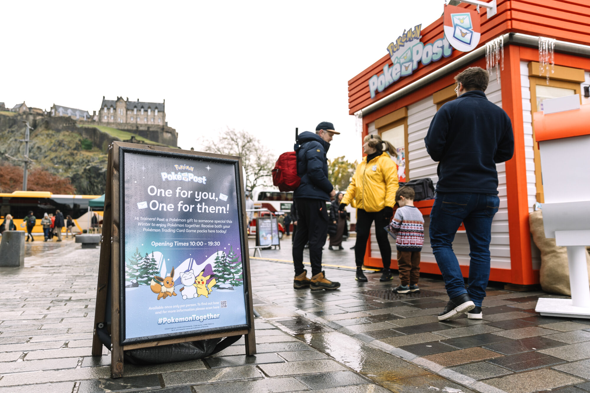 People stand near a Pokémon-themed kiosk and a sign promoting the Pokémon Poke Post on a wet city street. The sign features Pikachu and Eevee, with a castle visible in the background on a cloudy day.