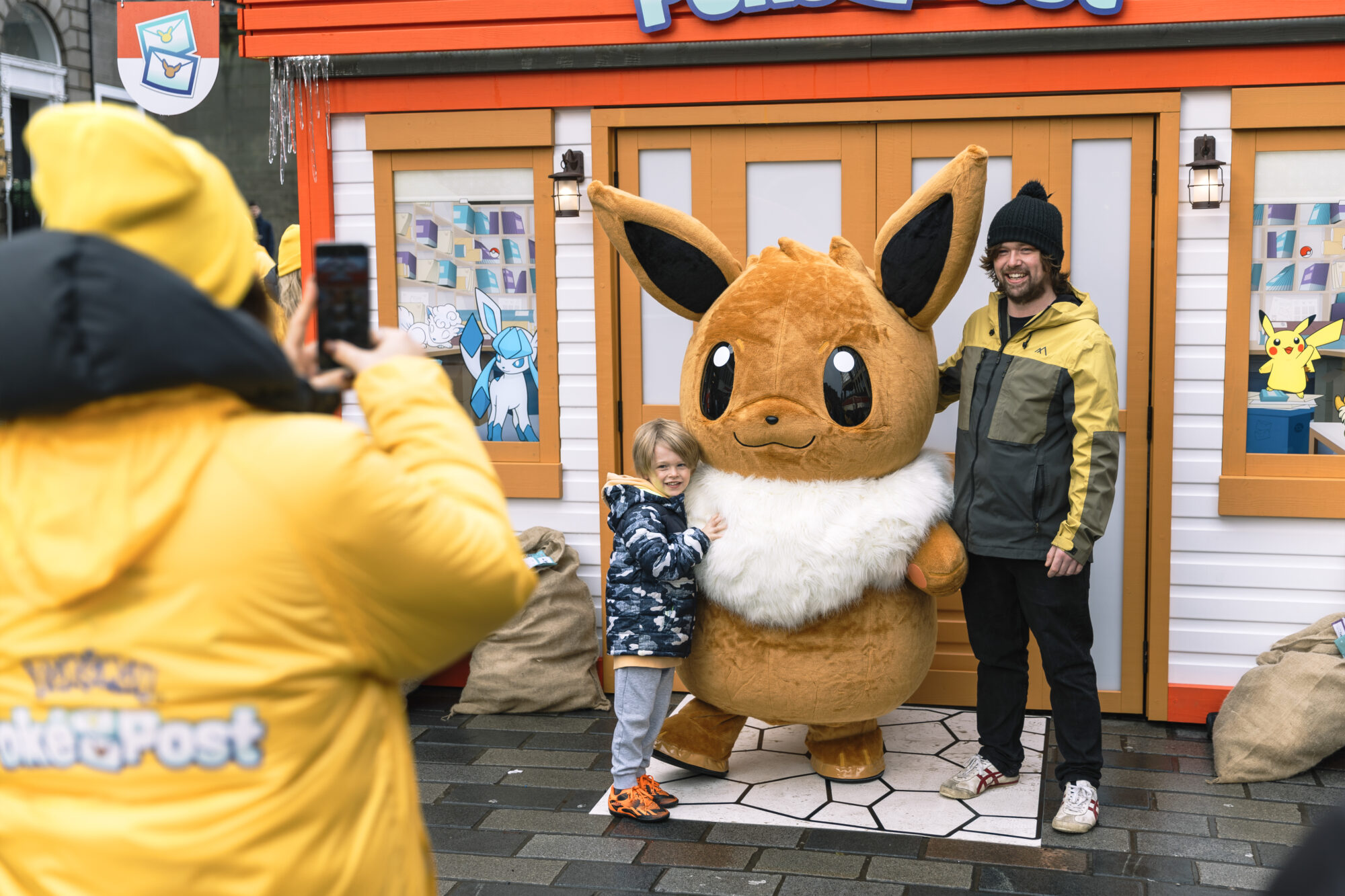A child and an adult pose with a large Eevee mascot outside a colorful Pokémon-themed booth, part of the Pokémon Poke Post, while someone in a yellow jacket takes their photo.