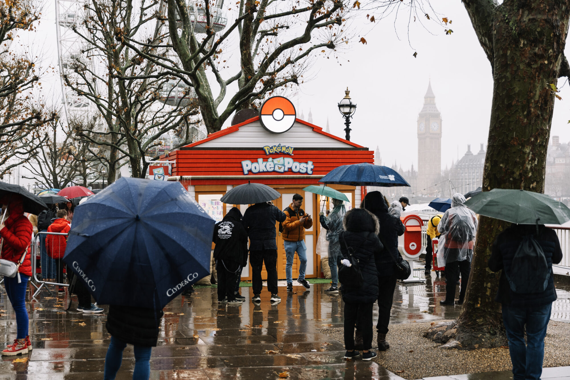 People with umbrellas stand in front of a Pokémon Poke Post booth on a rainy day in London. The booth features a red and white Poké Ball roof, with Big Ben visible in the background.