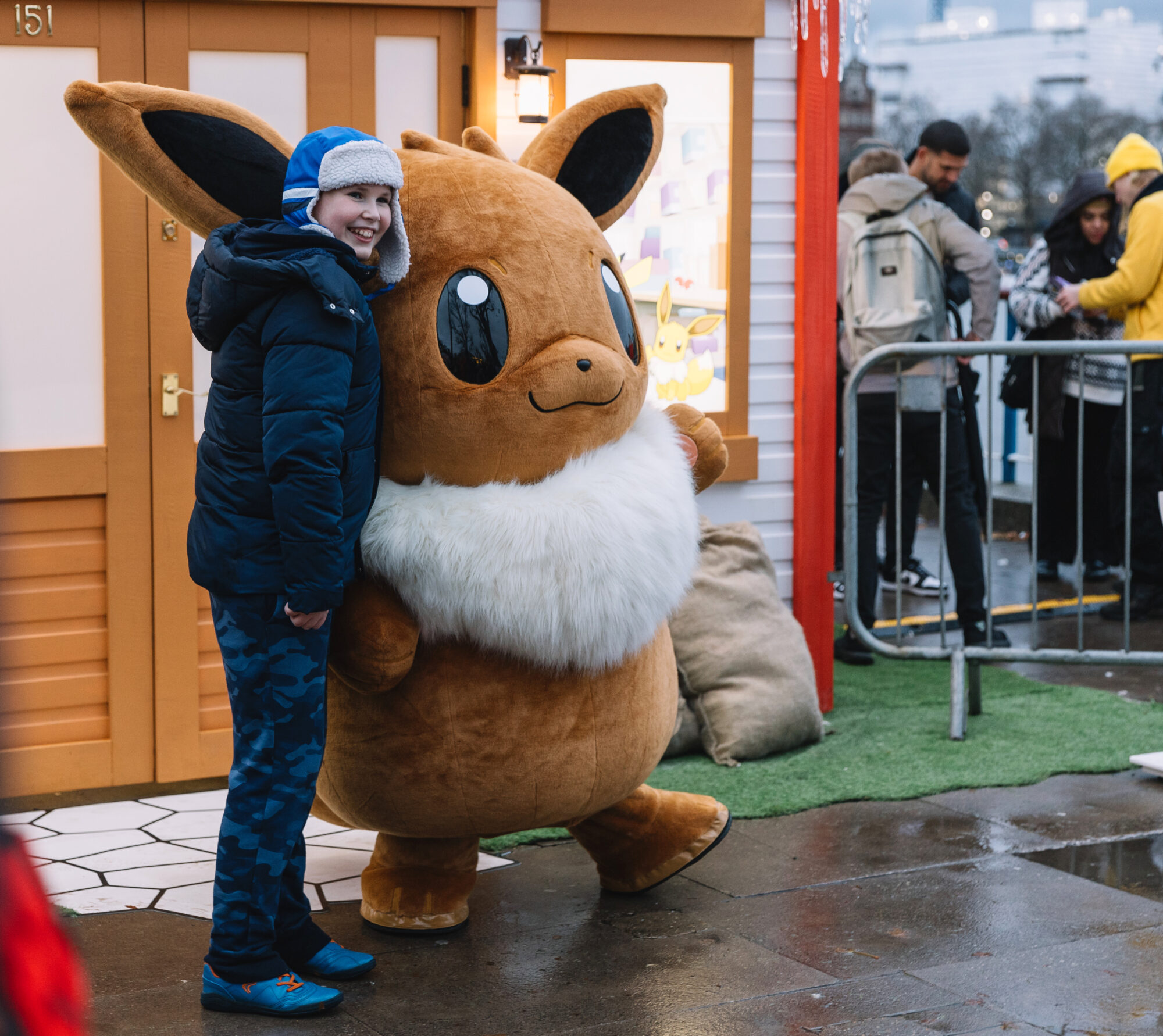 A smiling child in blue winter clothes stands next to a person in a large Eevee Pokémon costume outside a building, with other people, Pokémon Poke Post decorations, and festive touches visible in the background.