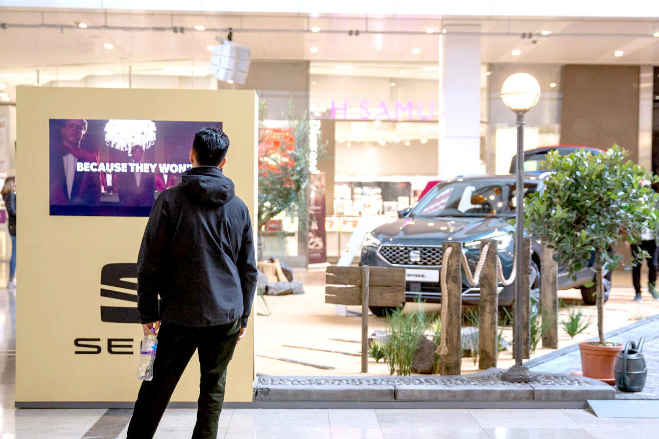 A person stands in a shopping mall, facing a SEAT car display with a screen reading BECAUSE THEY WON. A gray SUV is showcased behind decorative fencing and plants—a perfect scene for a SEAT case study. The person holds a drink bottle.