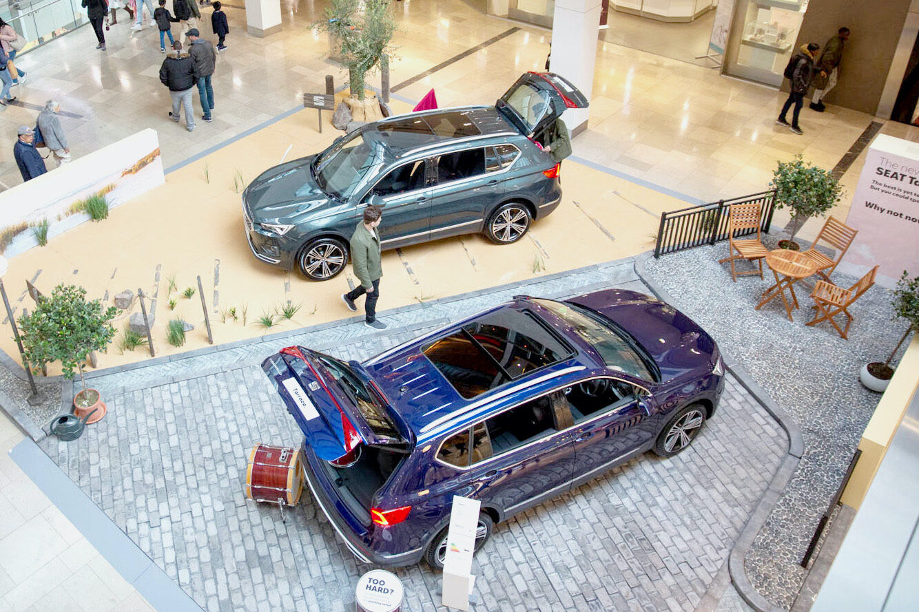 Two SUVs are displayed indoors in a shopping mall, with open doors and hatches. As part of the SEAT case study, people walk nearby, and there is a small patio setup with a table and chairs beside one of the cars.