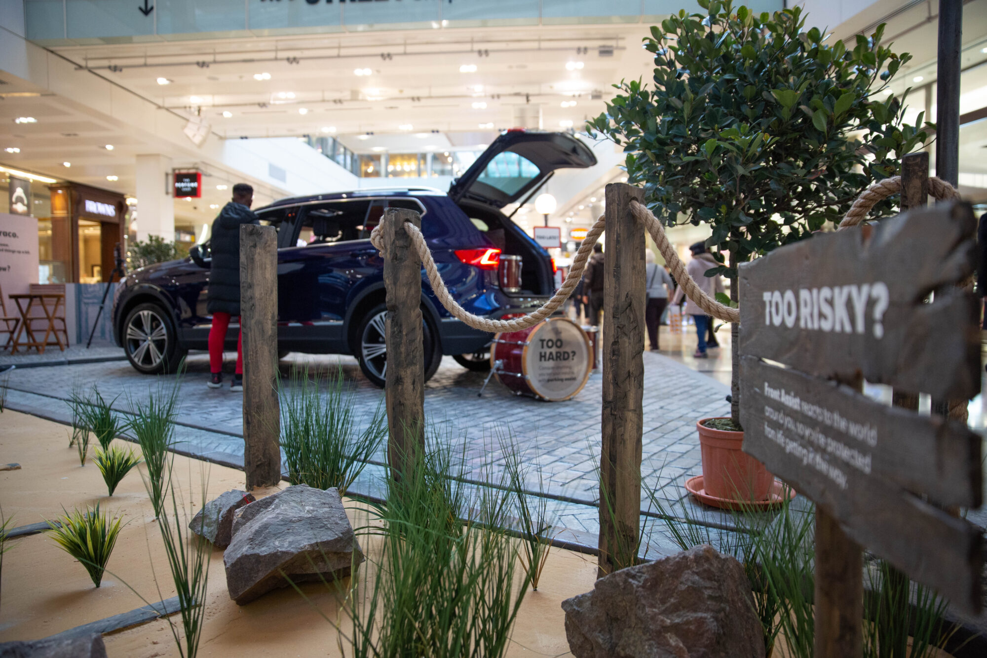 A blue SUV with its trunk open is displayed inside a shopping mall as part of a SEAT case study. People are standing nearby amid plants, rocks, and a wooden fence with a “TOO RISKY?” sign in the foreground.
