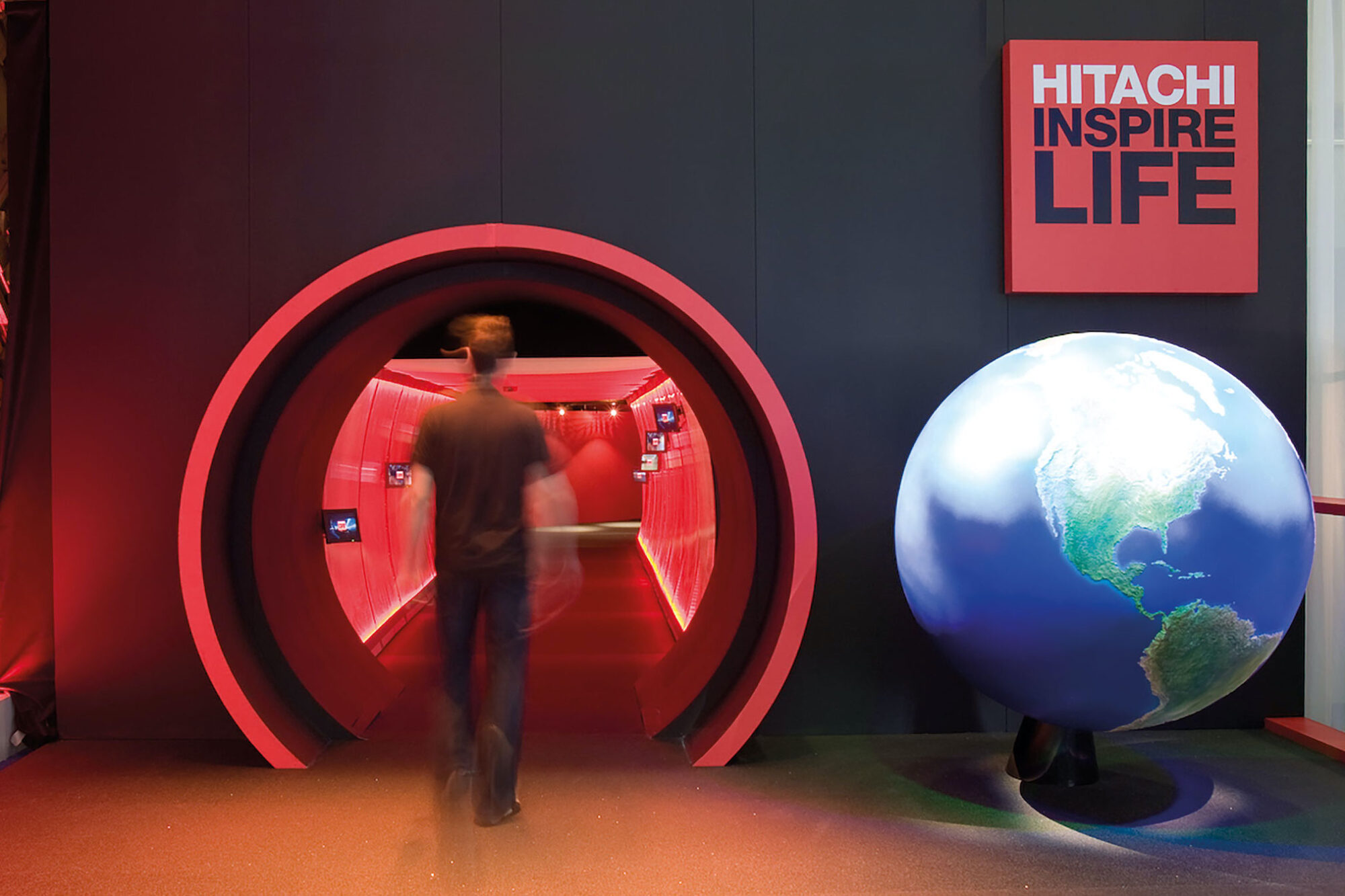 A person walks into a circular, red-lit tunnel beside a large globe displaying the Americas. A Hitachi Inspire Life sign glows on the wall, emphasizing innovation and inspiration.