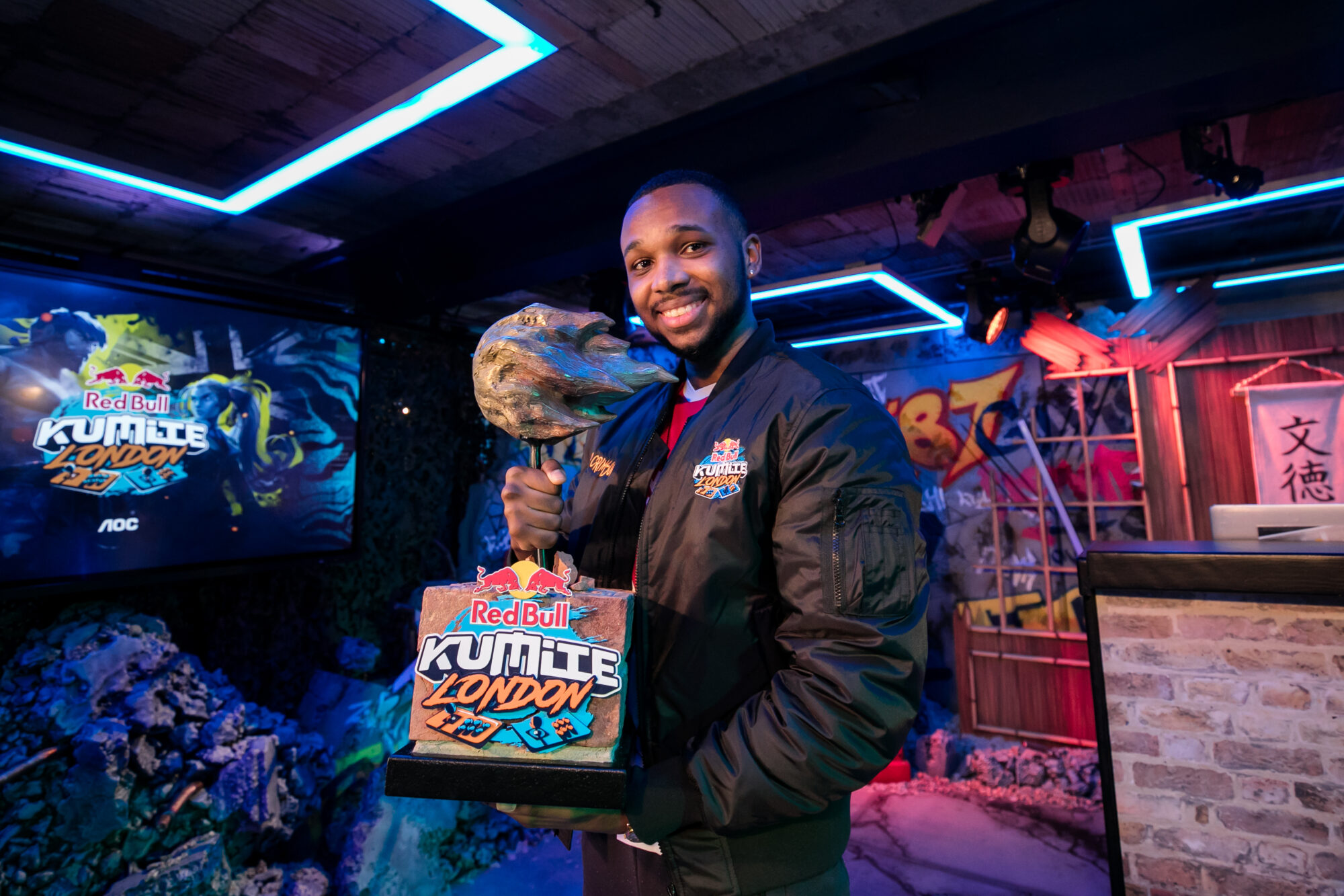 A smiling man in a Red Bull Kumite jacket holds a trophy shaped like a fist, with a sign reading “Red Bull Kumite London.” He stands in a neon-lit gaming venue featuring graffiti and the bold Red Bull Kumite logo in the background.