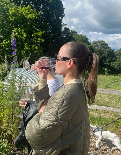 A woman from the marketing team wearing sunglasses and a green quilted jacket enjoys wine outdoors on a sunny day, with greenery, a wooden fence, and a small white dog on a leash in the background.