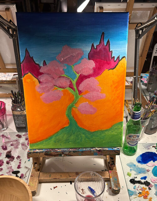 A colorful painting on an easel shows a pink-blossomed tree with orange hills and magenta mountains under a blue sky. Painting supplies, brushes, and a bottle of sparkling water await the marketing team on the cluttered table.