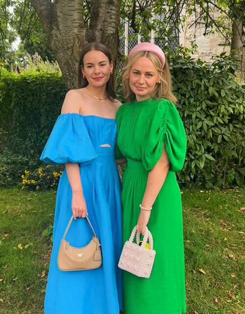 Two women, possibly from a marketing team, stand on grass before trees and bushes. One wears an off-shoulder blue dress with a tan handbag; the other dons a green dress with puffed sleeves, pink headband, and carries a small beaded bag.