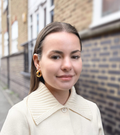A woman with straight brown hair, part of the marketing team, wears gold hoop earrings and a cream-colored coat as she stands outdoors in front of a brick wall, smiling softly at the camera.