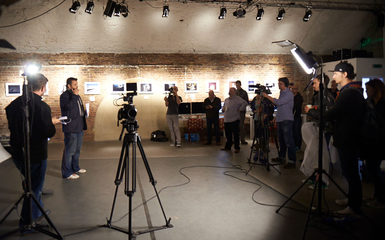 A group of people in a studio set with cameras, lights, and equipment films a man speaking in front of a brick wall with framed photos. Crew members observe and operate the cameras for this G-Tech case study production.