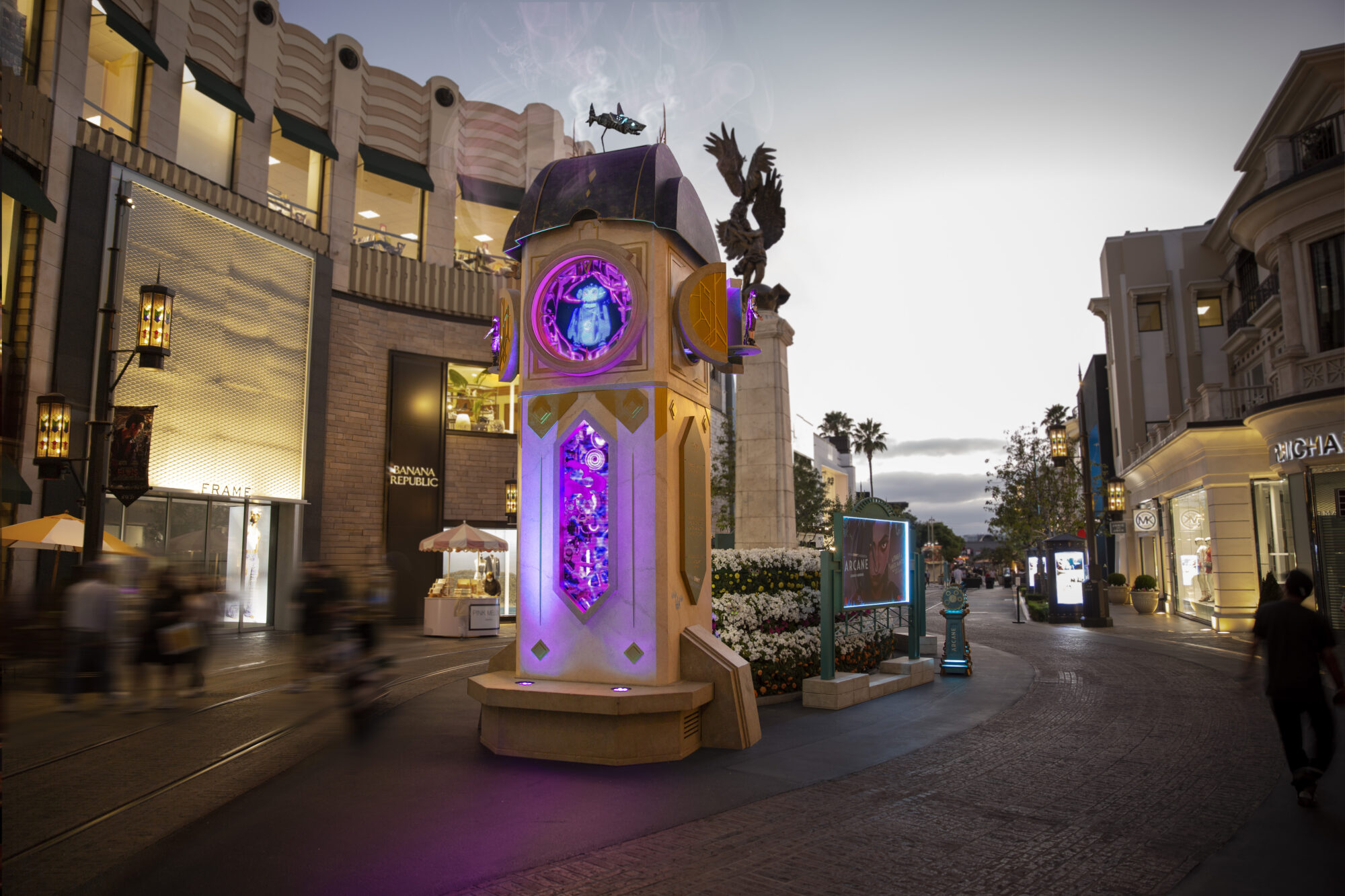 A large, ornate clock tower with glowing purple and blue lights stands in an outdoor shopping area at dusk, surrounded by shops and decorative flowerbeds—its magical glow reminiscent of the world of Arcane FYC Emmys. A few people walk nearby.