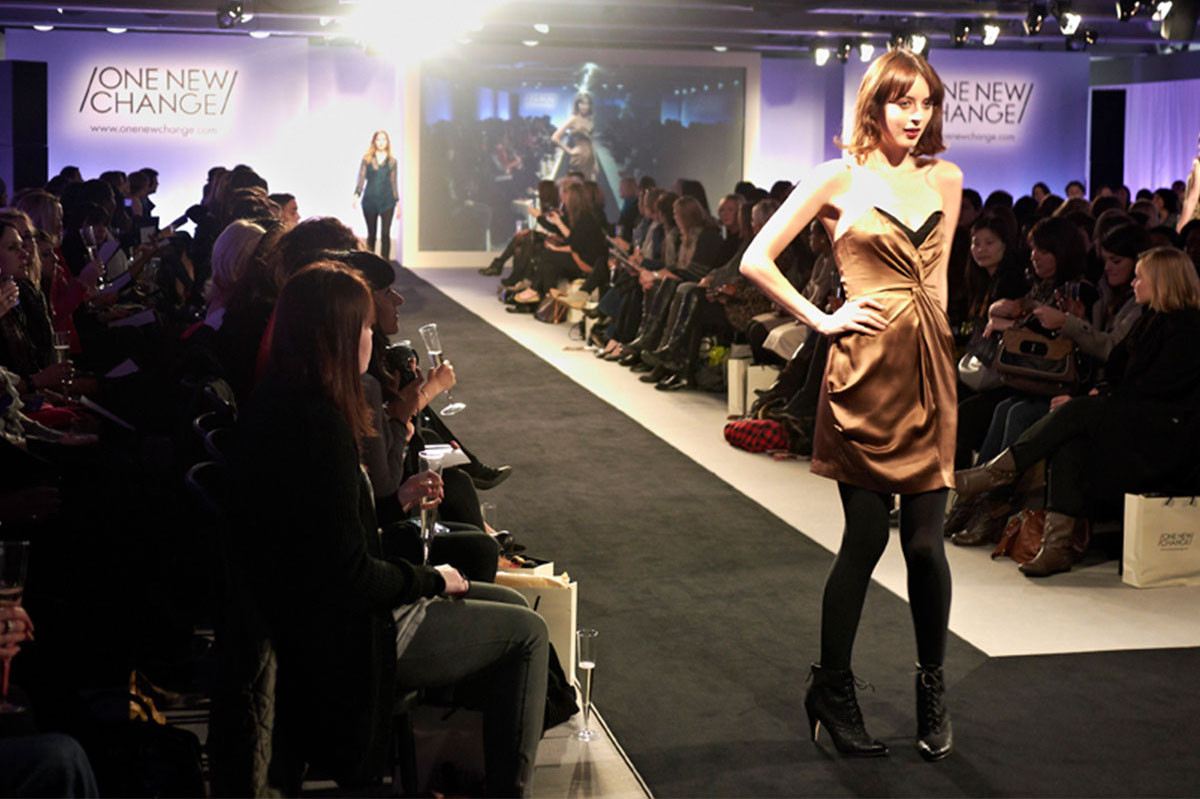 A model in a brown dress walks down a runway at a fashion show, with an audience on both sides—an atmosphere reminiscent of the Landsec case study, where bright lights illuminate the stage and a large screen dominates the background.