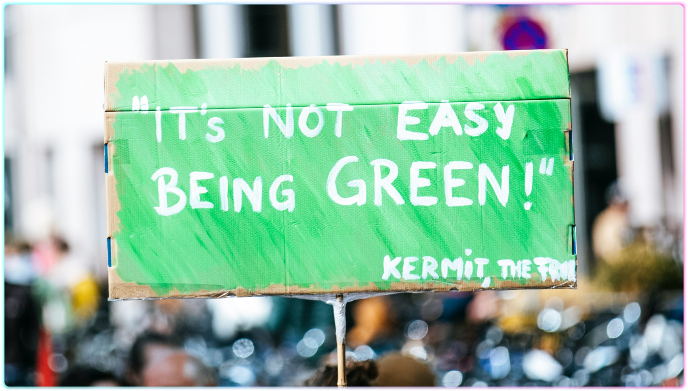 A cardboard sign painted green reads Its not easy being green! Kermit the Frog and is held up at an outdoor event focused on CSR and sustainability, with blurred people and bicycles in the background.