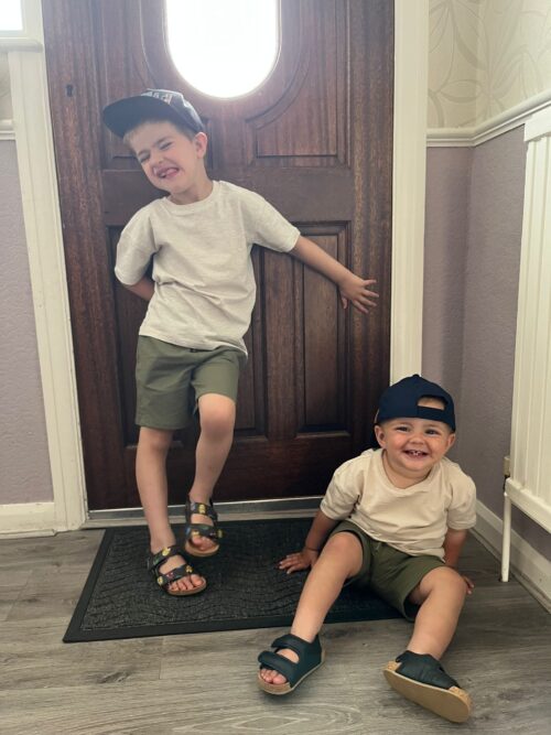 Two young boys in matching outfits—white t-shirts, green shorts, and dark sandals—smile playfully near a wooden door. Like a marketing team brainstorming ideas, one stands with a tilted cap while the other sits on the floor, both looking cheerful.
