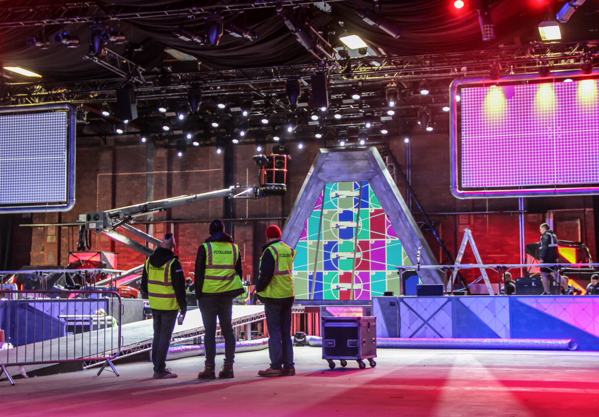 Three people in high-visibility vests stand in a studio, facing a colorful geometric set with bright lights and large screens for the Red Bull Home Ground event, while others work with production equipment in the background.