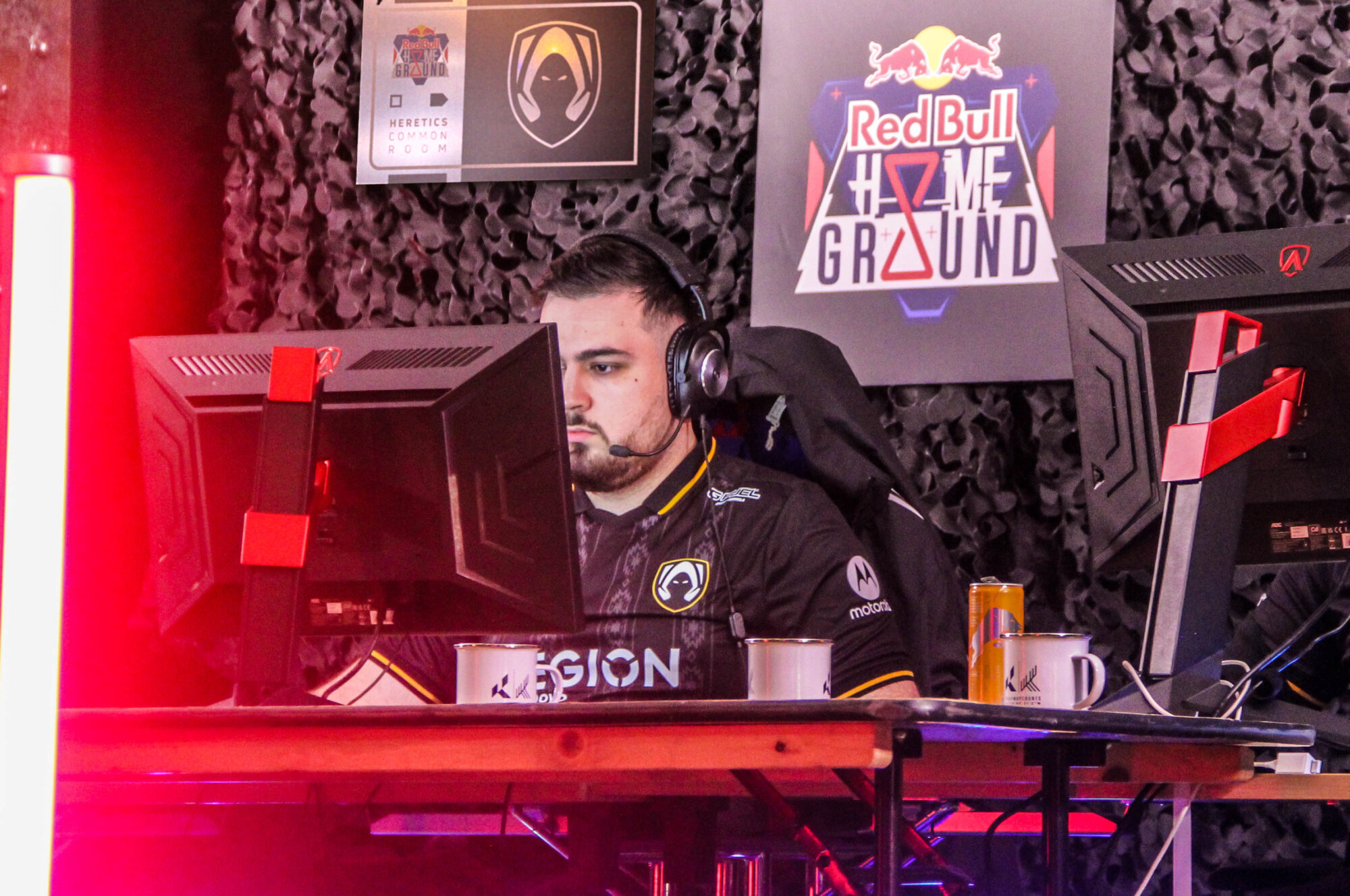 A focused esports player wearing a headset competes at a gaming setup with two monitors, mugs, and a glowing Red Bull Home Ground sign in the background, all illuminated by vibrant pink-red light.