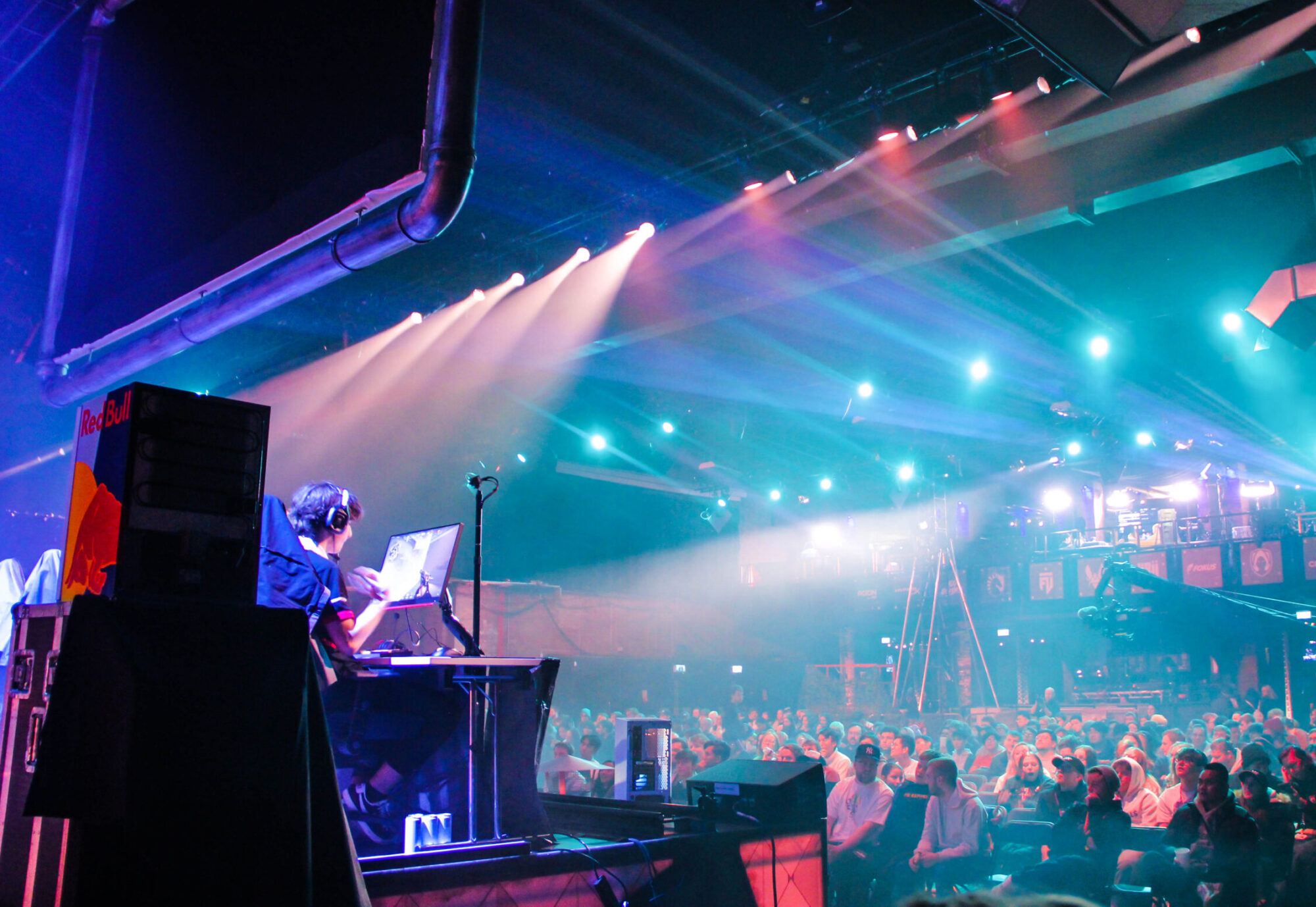 A DJ performs on stage under colorful lights in a large venue at the Red Bull Home Ground event, with a cheering crowd seated and standing in the background, creating a lively and energetic atmosphere.
