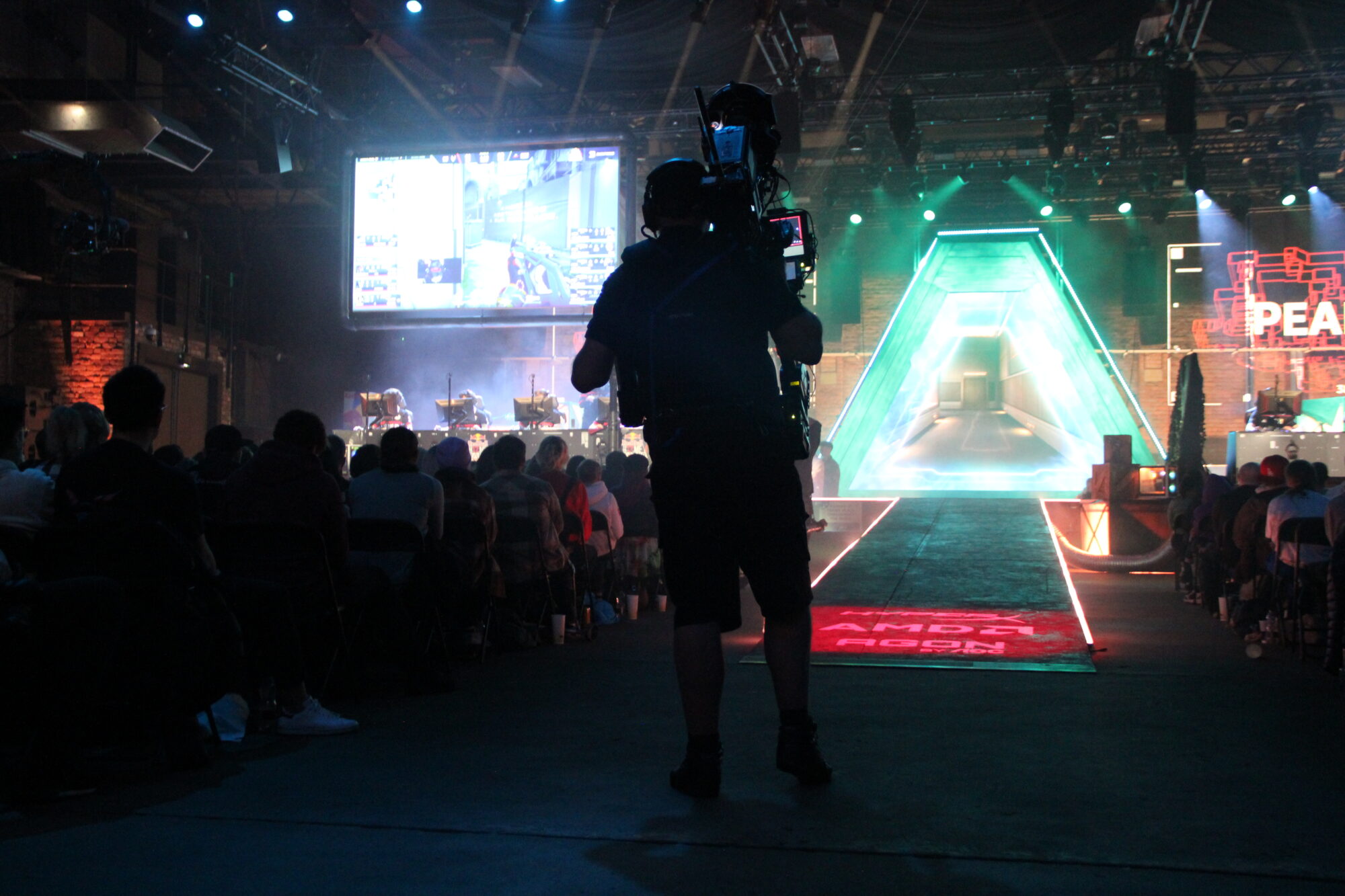 A camera operator films a live esports event inside a large, dimly lit arena. The audience faces a colorful, glowing stage inspired by Red Bull Home Ground, with screens and gaming stations shining under bright lights.