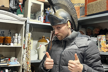 A man in a gray jacket gives two thumbs up while wearing a Roman-style helmet in a cluttered antique shop filled with various collectibles and vintage items.