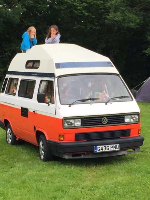 Two girls from the marketing team are looking out from a pop-up roof on a red and white Volkswagen camper van parked on grass, with trees and a tent in the background.