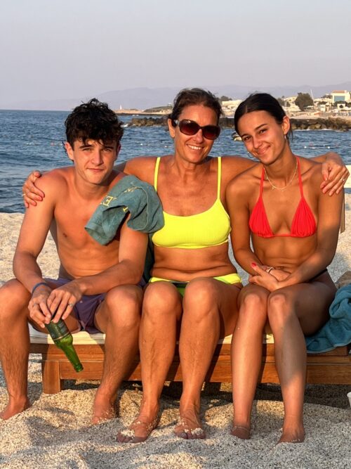 Three members of a marketing team sit closely together on a beach lounge chair, smiling at the camera. The woman in the center wears a yellow bikini, flanked by a young man and another woman in red, with the ocean in the background.