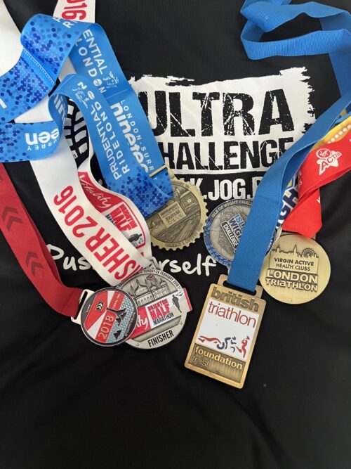 Five athletic medals with colorful ribbons are spread out on a black Ultra Challenge t-shirt, highlighting achievements in triathlons, marathons, and fitness challenges—perfect inspiration for any marketing team looking to celebrate success.