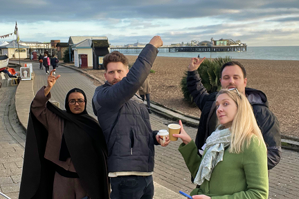 Four people stand on a seaside promenade holding coffee cups and pointing toward a distant pier. The beach and pier are in the background under a cloudy sky.