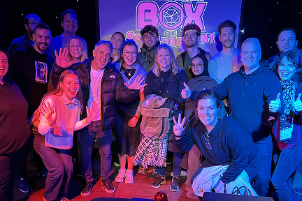 A cheerful group of people pose together on stage in front of a “Box of Entertainment” sign, smiling and waving at the camera under blue and purple lighting.
