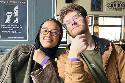 Two people sit side by side in a cozy indoor setting, smiling and showing purple wristbands. One wears glasses and a hijab, the other has curly hair and glasses. Posters and bright windows are visible in the background.