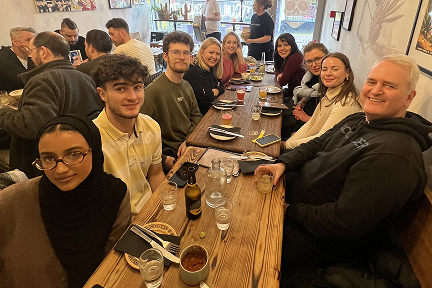 A group of eleven people sit around a long wooden table at a restaurant, smiling at the camera. The table is set with drinks, food, and utensils. Colorful art decorates the walls behind them.