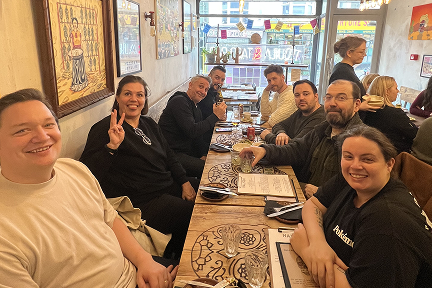 A group of eight people sit around a long restaurant table, smiling and posing for the camera. One person flashes a peace sign. Menus, drinks, and cutlery are on the table; the atmosphere is casual and lively.