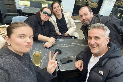 Five people sit around a gray outdoor table, smiling and posing for a selfie. One person flashes a peace sign, another sticks out their tongue. There’s a drink and a phone on the table.