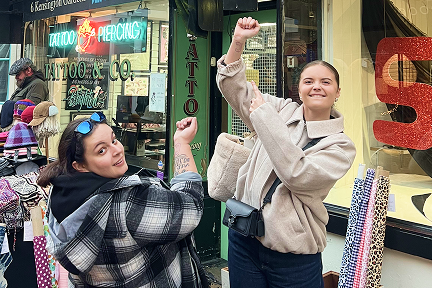 Two women stand outside a tattoo and piercing shop, smiling and proudly showing off fresh tattoos on their forearms. One wears a plaid jacket, the other a beige coat. Rolls of colorful wrapping paper are visible nearby.