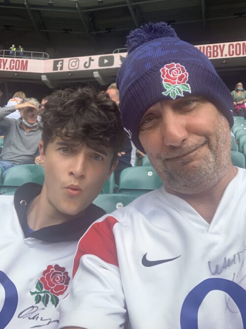 Two people from the marketing team wearing England rugby shirts, and one in a navy hat with a red rose logo, smile and pose for a selfie in a stadium filled with rows of seats and spectators in the background.