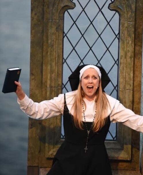 A woman dressed as a nun holds a book in one hand and sings with arms outstretched, as if leading a marketing team, in front of a stained glass window, wearing a black and white habit and a rosary.