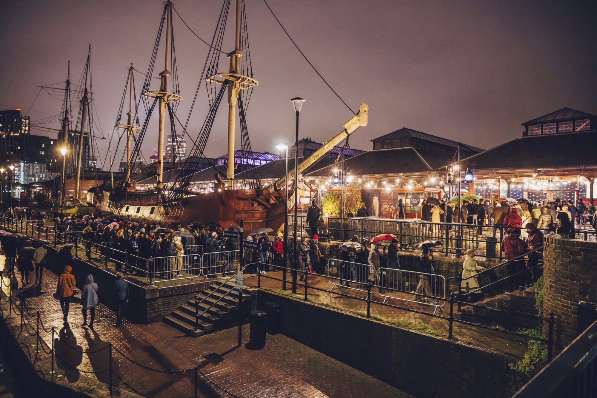 A large crowd waits in line at night beside a historic tall ship and a lit brick building, with string lights and umbrellas visible on a rainy evening during the Waitrose Winter Festival.