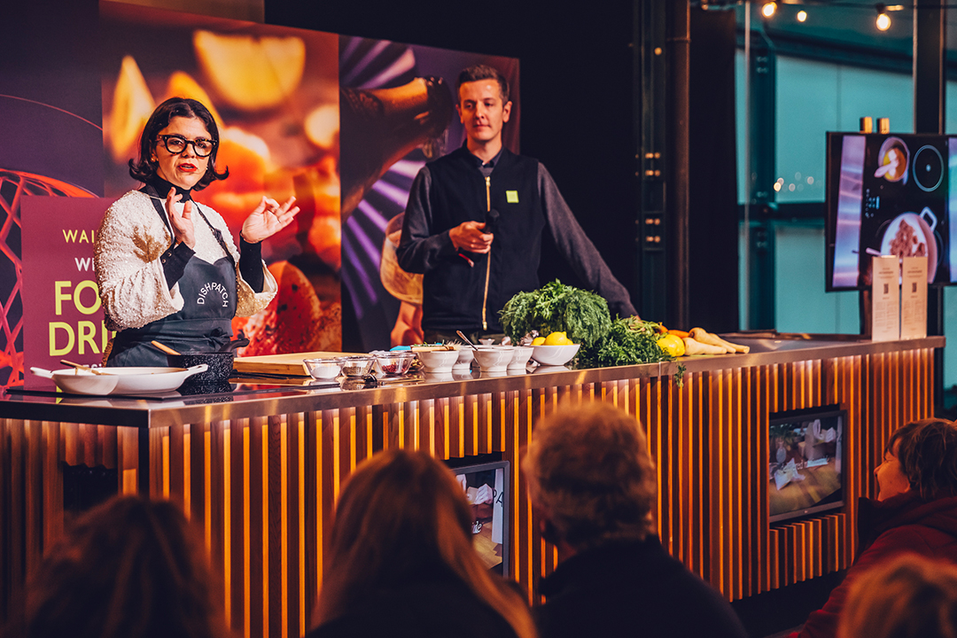 A woman in an apron speaks to an audience during a Waitrose Winter Festival cooking demonstration, with a man standing beside her behind a counter filled with fresh vegetables, lemons, and kitchen utensils.