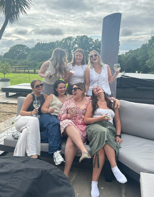 Seven women from a marketing team sit and stand on outdoor sofas, laughing and smiling while holding drinks. They appear to be enjoying a relaxed, sunny day together, with trees and greenery in the background.