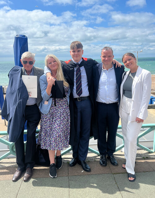 Five people stand together outdoors by the seaside on a sunny day, smiling. The central figure wears a graduation gown and cap, flanked by proud marketing team members dressed in smart clothing. A blue sky and ocean are in the background.