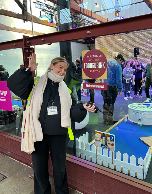 A smiling member of the marketing team in a scarf and coat stands indoors, gesturing toward a glass door with a Waitrose Winter Food & Drink Festival sign. People and a mini-golf setup are visible inside the venue.