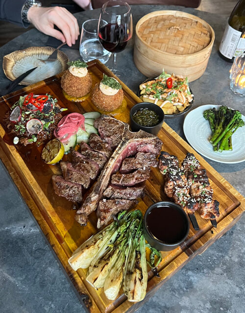 A wooden board with sliced grilled steak, grilled spring onions, two sauces, pickled vegetables, and two stuffed balls—perfect for sharing with your marketing team. Nearby are grilled broccolini, salad, a bamboo steamer basket, and glasses of water and red wine.
