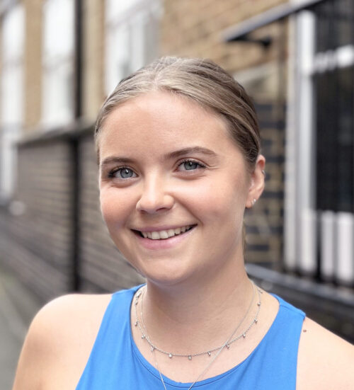 A young woman with light skin and blonde hair tied back smiles at the camera. She is wearing a blue top and delicate necklaces, standing outdoors next to a brick building—ready for another day with her marketing team.