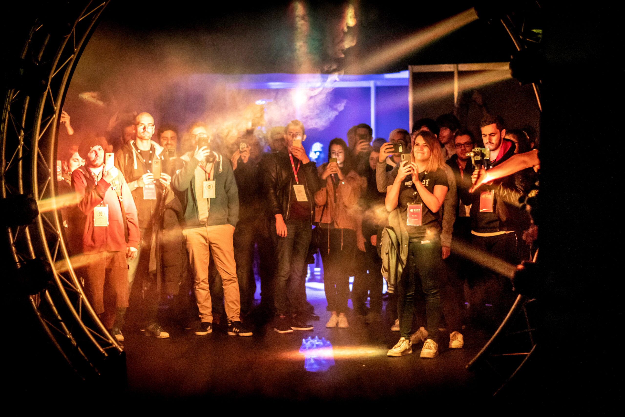 A crowd of people, many holding up phones to take photos or videos, stands in a dimly lit, smoky environment facing the Riot Games L10 circular structure with bright lights, creating an energetic and immersive atmosphere.
