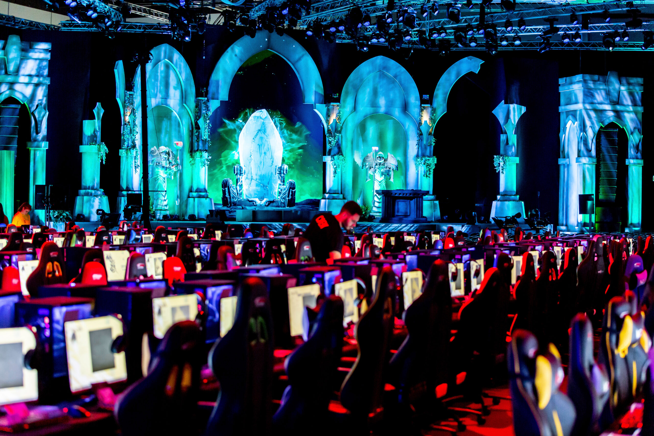 A large, dimly lit Riot Games L10 event features rows of empty computer stations and a fantasy-themed stage in the background, decorated with glowing blue-green arches and statues, creating an immersive atmosphere.