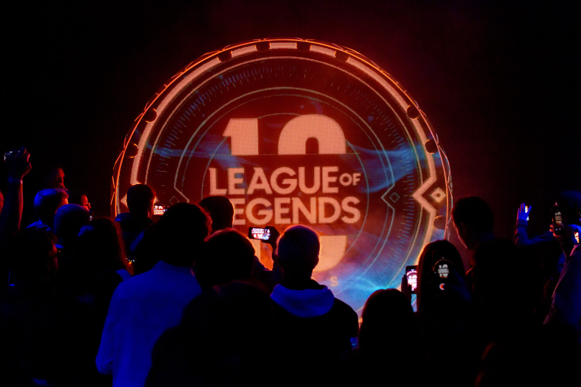 A crowd watches and records a glowing, circular Riot Games L10 display celebrating the 10th anniversary of League of Legends, with the words LEAGUE OF LEGENDS and 10 prominently shown.