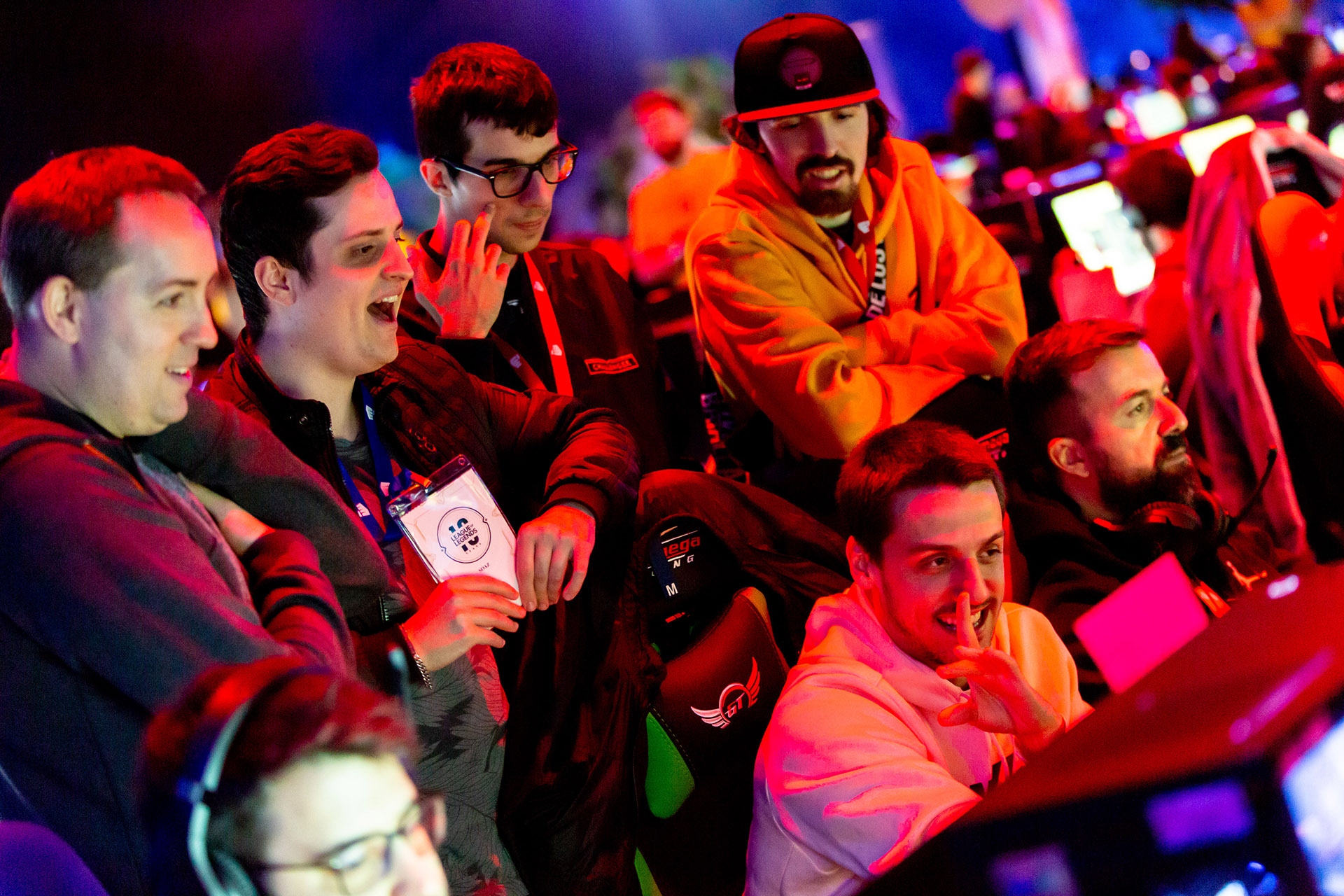 A group of enthusiastic young men gather around gaming monitors at a Riot Games L10 esports event, smiling and reacting excitedly to what they see on the screens in a vibrant, colorful setting.