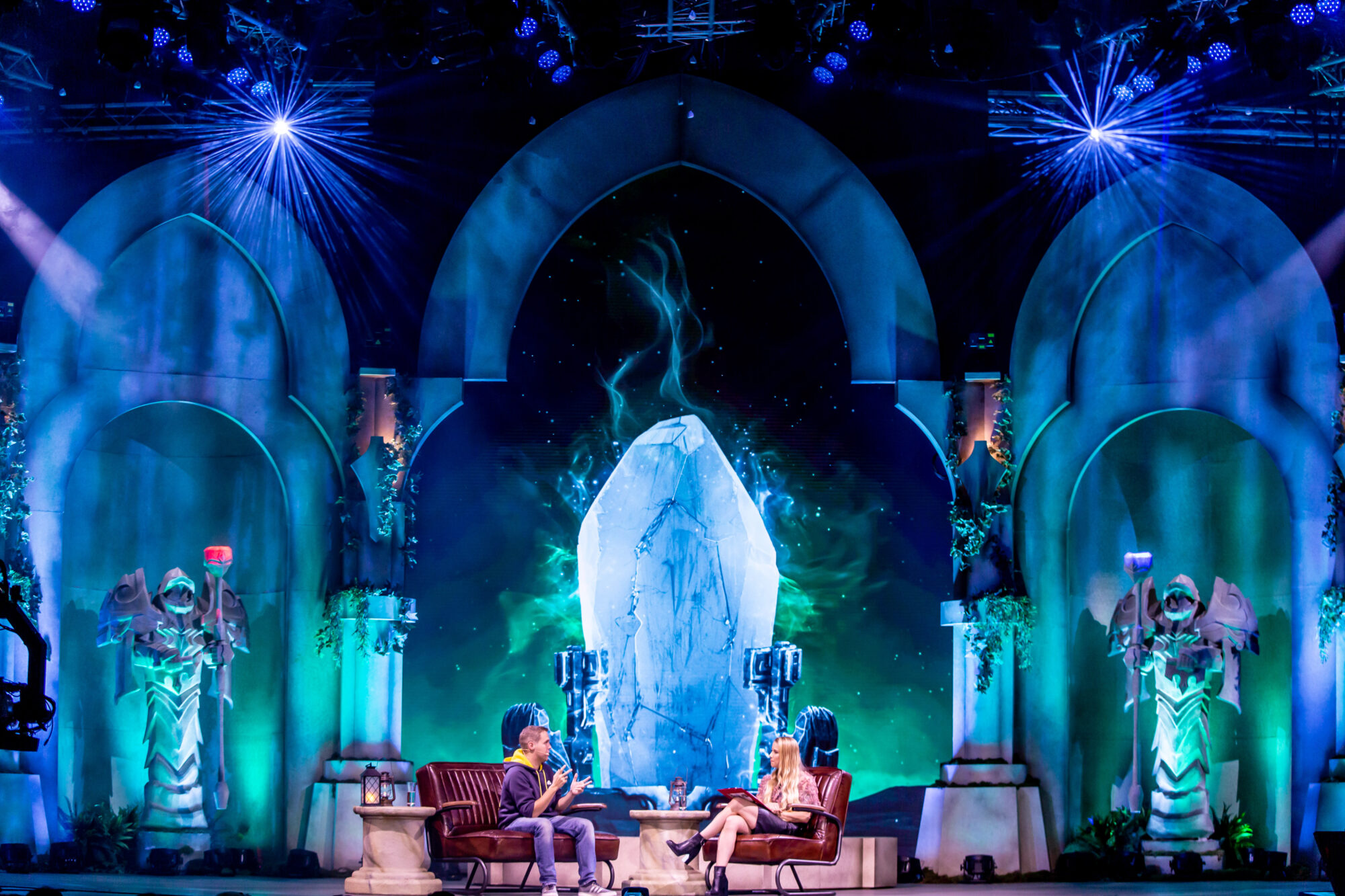 Two people sit and talk onstage at Riot Games L10 in front of an elaborate fantasy-themed backdrop featuring a glowing crystal, arched structures, and mystical statues, with dramatic blue and green lighting creating a magical atmosphere.