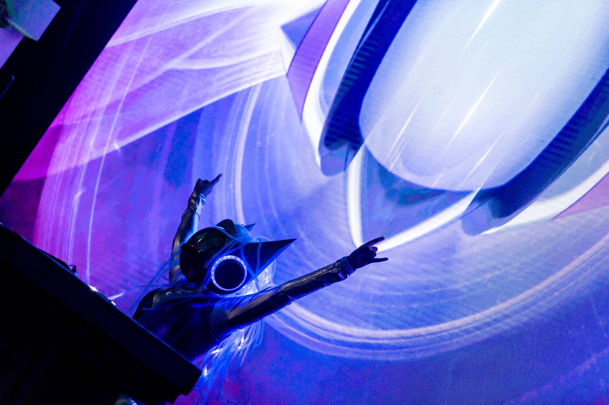 A person wearing a futuristic helmet with cat ears and glowing lights raises their arms in front of a vibrant, abstract digital screen inspired by Riot Games L10, awash in striking purple and blue hues.