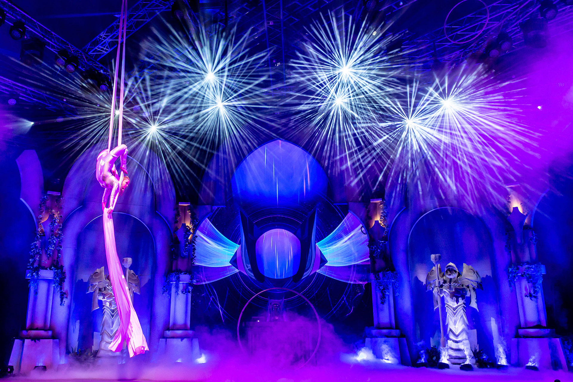 A performer in a pink costume does aerial silks on a dramatic stage with purple lighting and fog, as bright spotlights illuminate ornate set pieces inspired by Riot Games L10, surrounded by figures in elaborate costumes.