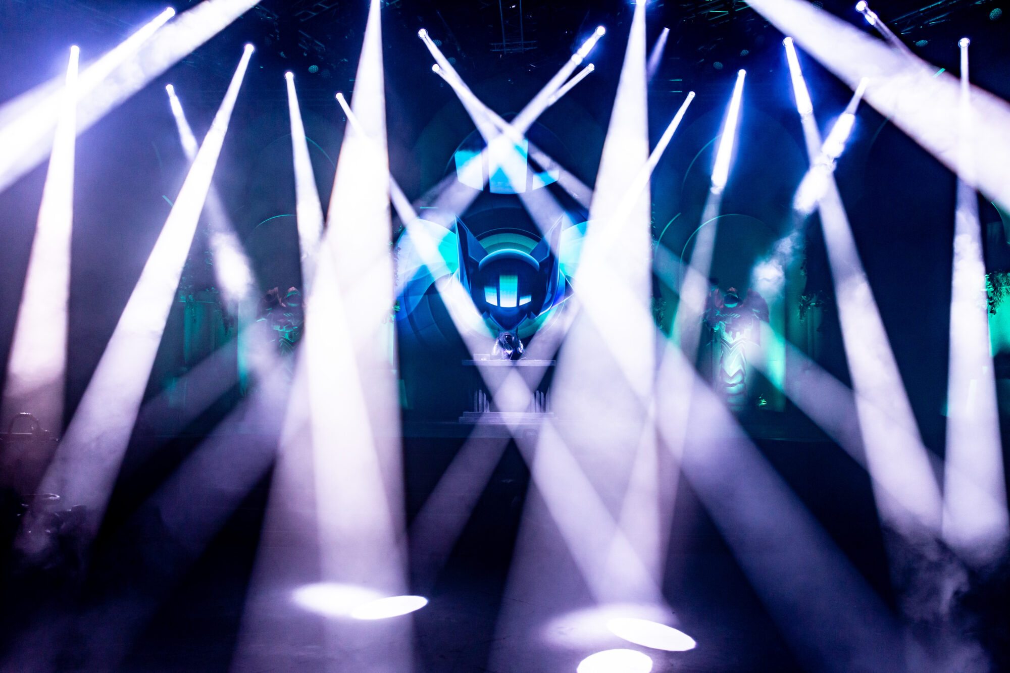 A stage filled with bright white and blue spotlights shines through smoke, creating dramatic beams of light in every direction. At the center, a DJ at the console energizes the crowd with colorful lighting effects at Riot Games L10.